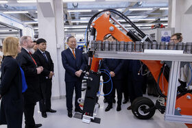 En grupp människor i formell klädsel observerar maskiner i en industriell miljö. Maskinen har ljusa orangea komponenter.