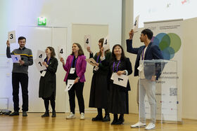 Six people holding numbered signs at an event, standing in front of podium and screen