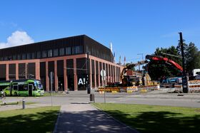 En röd och gul byggarbetsplats med en grön spårvagn framför en modern tegelbyggnad under en klarblå himmel.