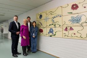 Four people stand in front of a large artwork featuring flowers and leaves displayed on a white brick wall.