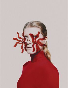 A person with long blonde hair wearing a red turtleneck and red abstract earrings.