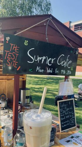 A summer cafe with a blackboard sign displaying hours and specials. A creamy drink is in the foreground.