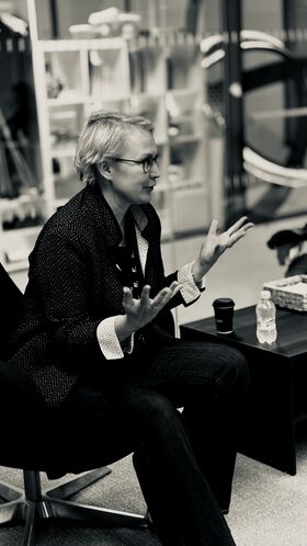 A person gesturing with both hands while sitting on a chair. Bottled water and a coffee cup are on the table beside them.
