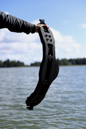 A person holds a black foam aqua jogging belt with holes, against a background of a lake and trees under a blue sky.