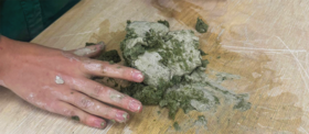 A person's hand is kneading grey and green clay on a wooden surface. The clay mixture is messy and spread out.