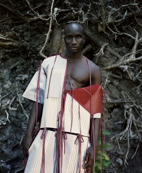 A person wearing an asymmetrical outfit with red straps stands in front of exposed tree roots.