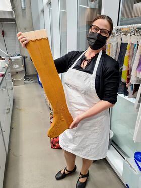 Henkilö valkoisessa essussa pitää pitkää, sinapinväristä neulottua sukkaa työpajassa, jossa on erilaisia kankaita ja työkaluja.