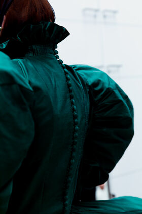 Lähikuva henkilöstä, jolla on tummanvihreä mekko, korkea röyhelökaulus ja kangaspäällysteiset napit selässä.