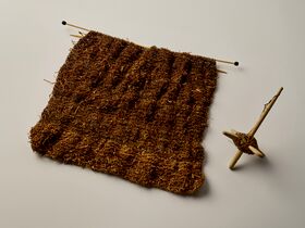 A brown knitted piece on needles with a spindle on a light surface.