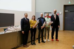 Viisi ihmistä seisoo sisällä pitäen kimppuja. Vieressä on pöytä, jossa on lisää kimppuja. Näkyvissä on ovi ja näyttö.