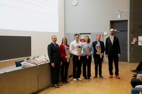 Kuusi ihmistä seisoo huoneessa, viidellä on kukkakimput. Taustalla näkyy esitysnäyttö ja kello.