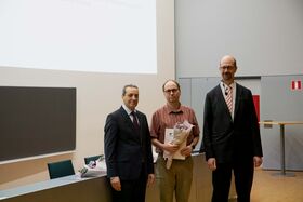 Kolme miestä seisoo huoneessa, yksi pitää kukkia ja todistusta. Näkyvissä on esitysnäyttö ja liitutaulu.