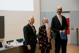 Kolme henkilöä seisoo sisällä juhlapuvuissa. Yksi pitää kukkakimppua, toinen kansiota. Näkyvissä tuoleja ja pöytiä.