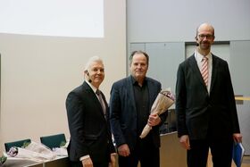 Kolme miestä puvuissa seisoo sisällä. Yksi pitää kukkakimppua, toinen asiakirjaa. Näkyvissä tuoleja ja pöytiä.