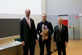 Kolme miestä puvuissa seisoo sisätiloissa, yksi pitää kukkakimppua. Taustalla näkyy pöytä ja punainen ovi.