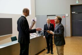 Mies ojentaa kukkakimpun toiselle miehelle luentosalissa. Kolmas mies seisoo taustalla.