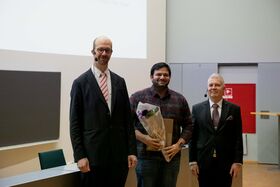 Kolme miestä seisoo sisällä, yksi pitää kukkakimppua. He ovat pukeutuneet muodollisesti, taustalla esitysnäyttö.