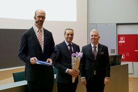 Kolme miestä puvuissa seisoo huoneessa. Yksi pitää kukkakimppua, toinen asiakirjaa. Punainen palosammutin näkyy.