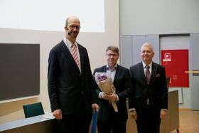 Kolme miestä puvuissa seisoo sisällä, yksi pitää kukkakimppua. Taulut ja punainen paloturvallisuuslaatikko näkyvät.