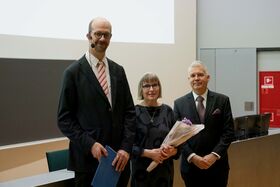 Kolme ihmistä seisoo sisätiloissa, yksi pitää kukkakimppua. He ovat pukeutuneet muodollisesti pukuihin ja mekkoon.