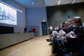 Luentosali, jossa esitys metsäkoneista. Yleisön jäsenillä on kukkakimppuja.