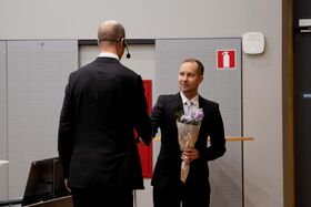 Kaksi miestä puvuissa kättelee sisätiloissa. Toinen pitää kukkakimppua. Seinällä näkyy palosammuttimen merkki.