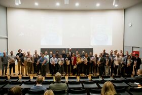 Ryhmä ihmisiä seisoo luentosalissa, pitäen todistuksia ja kukkia. Suuri näyttö näkyy heidän takanaan.