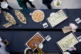 Buffetpöytä, jossa on erilaisia ruokia, kuten salaatteja, leivonnaisia ja lämpimiä ruokia. Ihmiset ottavat ruokaa.