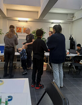 People standing and sitting in a room with tables and chairs, engaging in various activities. A child is pointing at something.