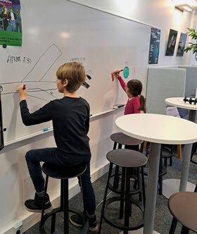 Two children drawing on a whiteboard in a classroom. One draws an airplane, the other colours a shape. Posters are on the wall.