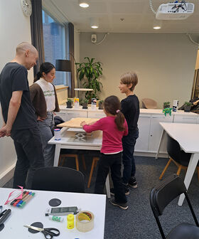Four people are gathered around a table with craft supplies in a room with a projector and a plant.