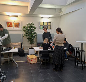 People are gathered in a modern room with tables, chairs, and a potted plant. Artworks are displayed on the wall.