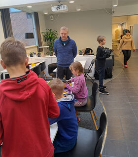 Children and adults in a room with tables and chairs. Some children are playing with toys on a table.