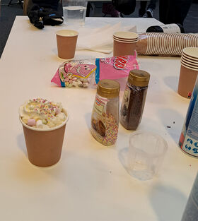A table with a hot drink topped with whipped cream and sprinkles, surrounded by cups, toppings, and a bag of marshmallows.