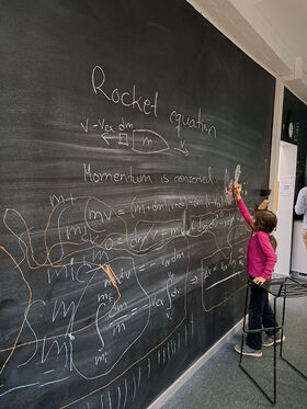 A child in a pink shirt writes on a large blackboard filled with rocket equations and diagrams in a classroom.