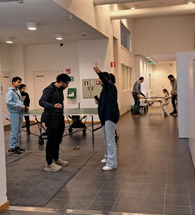 People playing table tennis indoors. Others are standing and talking. A child is playing at a table in the background.