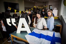 Group of people in a room with a large letter 'A' and quotation marks on a table, and a Finnish flag with signatures.