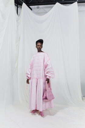 Henkilö pukeutunut pitkään vaaleanpunaiseen mekkoon, jossa on pussihihat, ja pitää vaaleanpunaista käsilaukkua. Valkoinen tausta.