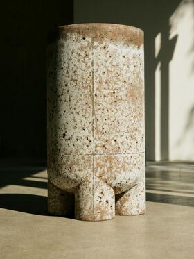 A cylindrical ceramic vase with a speckled brown and white pattern, standing on a concrete floor in sunlight.