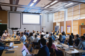 A classroom with students seated at tables, listening to a presentation. A projector screen displays an outline.