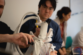 A person adjusts a white robotic arm with multiple joints and cables in a room with other people.