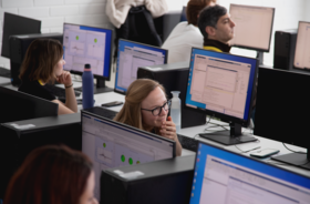 People working on computers in a classroom setting, with various screens displaying data and graphs.