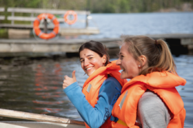 Two people wearing orange life jackets on a boat near a dock. One person is giving a thumbs up.