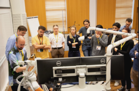 A group of people watching a demonstration involving a robotic arm in a classroom setting.