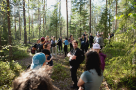 A group of people standing on a forest path surrounded by tall trees and greenery, some wearing backpacks.
