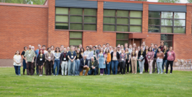 A group of people posing outside a brick building with large windows. They are standing on a grassy area.
