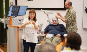 A medical demonstration with a seated person wearing electrodes, surrounded by equipment and two people explaining the process.