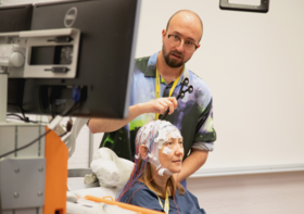 A person wearing a colourful shirt adjusts electrodes on another person's head in a lab setting.