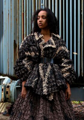 A person wearing a textured, oversized jacket with intricate patterns and a wide belt, standing in front of a corrugated metal wall.