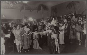 Vanha valokuva naamiaisista, joissa ihmiset ovat pukeutuneet hienoihin asuihin ja naamioihin. Esiintyjä tanssii etualalla.